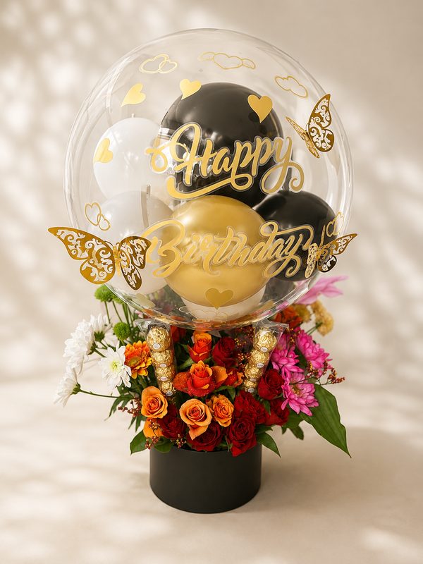 Bubble-Ballon mit goldenem Happy-Birthday-Schriftzug und goldenen Schmetterlingen, gefüllt mit schwarzen, weißen und goldenen Ballons, auf einer schwarzen Box mit frischen Rosen, Chrysanthemen und Ferrero Rocher