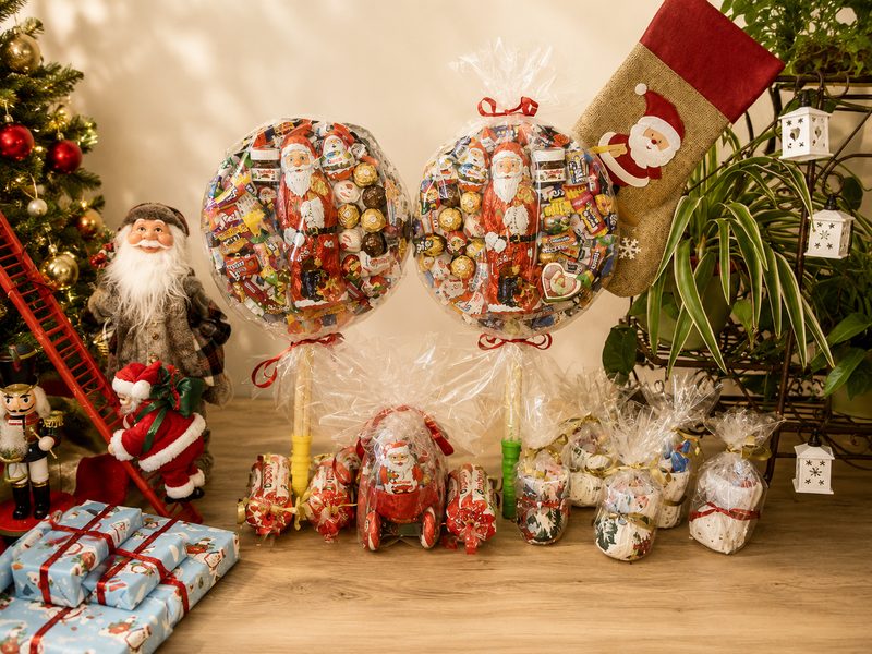 Zwei große Süßigkeitenkugeln am Stiel mit Schokoladen-Weihnachtsmännern, dazu kleine verpackte Präsente vor geschmücktem Christbaum und Nikolausstiefel